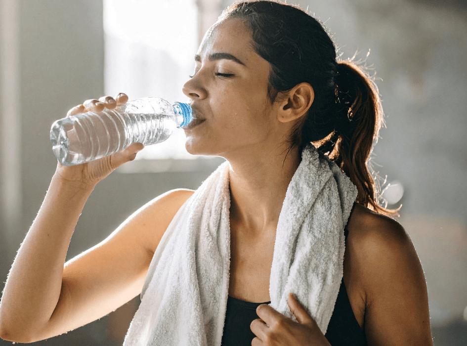 Mladá žena v športovom oblečení s uterákom okolo krku pije vodu z plastovej fľaše.