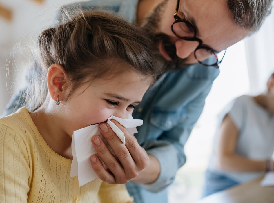 dieťa smrká do papierovej vreckovky, pričom mu otec pomáha pridržať vreckovku pri nose