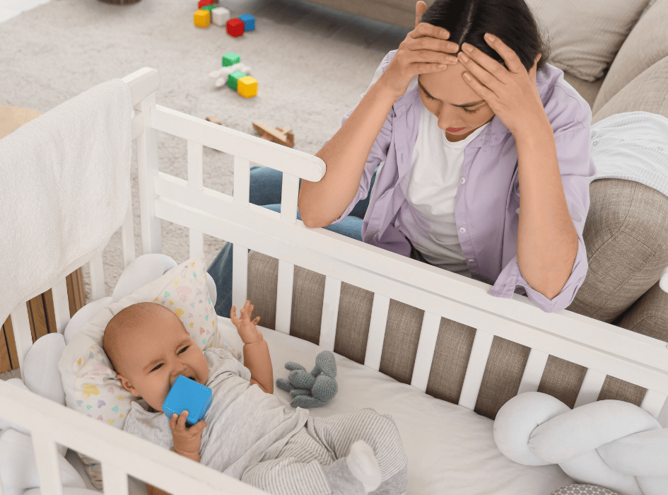 Unavená mama pri postieľke s bábätkom – stres, nedostatok spánku a psychická záťaž po pôrode.